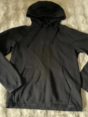 Svart hoodie från EA7 Emporio Armani - Snygg svart hoodie från EA7 Emporio Armani med broderad logga på bröstet. Klassisk modell med huva, dragsnören och stor magficka. Tillverkad i mjukt bomullsmaterial som är skönt mot huden. Perfekt för en avslappnad och trendig stil.