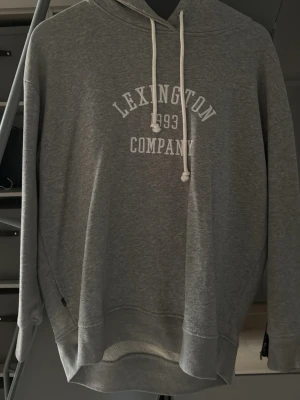 Grå hoodie från Lexington Company - Snygg grå hoodie från Lexington Company med vit text och årtal framtill. Klassisk modell med huva och vita dragsnören, samt ribbade muddar vid ärmslut och nederkant. Perfekt för en avslappnad och stilren look.