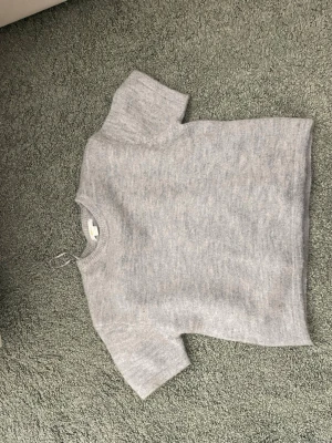 Ljusgrå stickad t-shirt - En stilren ljusgrå stickad t-shirt med korta ärmar och rund halsringning. Tröjan har en rak passform och är tillverkad i ett mjukt, melerat garn som ger en avslappnad look. Perfekt för lager-på-lager och enkel att matcha med andra plagg.
