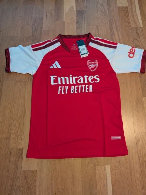 26/27 Arsenal matchtröja Adidas röd/vit. S.m storlek - Helt ny Arsenal matchtröja från Adidas i rött med vita ärmar och svarta detaljer. Tröjan har klubbmärke och sponsortryck på bröstet samt Adidas-logga. Tillverkad i lätt och ventilerande material, perfekt för fotbollsträning eller match.