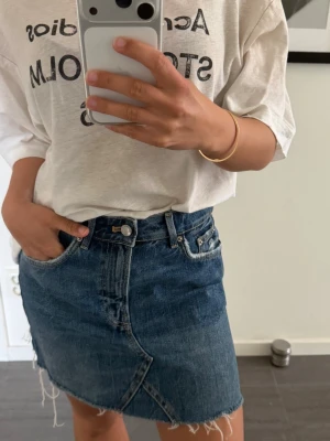 Blå jeanskjol från Zara Strl S - Snygg blå jeanskjol från Old Habits med råa kanter och klassisk femficksdesign. Kjolen är kort och har en omlottliknande detalj framtill. Perfekt för dig som gillar en avslappnad och trendig stil. 