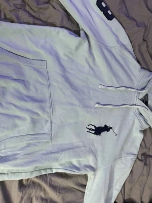 Ljus blå hoodie från Polo Ralph Lauren - Snygg ljus blå hoodie från Polo Ralph Lauren med stor mörkblå polospelare broderad på bröstet och siffran 3 på ena ärmen. Klassisk känguruficka framtill och dragsko i huvan. Tillverkad i mjukt bomullsmaterial som är skönt att ha på sig.