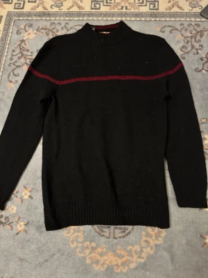 Svart stickad tröja med röd detalj - Svart stickad tröja med lång ärm och en tunn röd rand över bröstet och armarna. Tröjan har ribbstickade muddar vid ärmslut och nederkant samt en liten ståkrage. Perfekt för dig som gillar enkel men snygg design.