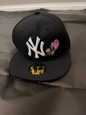 Svart New Era NY keps med rosbrodyr - Snygg svart 59FIFTY keps från New Era med klassisk NY-logga i vitt och broderade rosa rosor på framsidan. Platt skärm med rosa undersida ger en unik touch. Perfekt för dig som vill sticka ut med din stil.