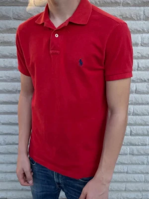 Röd Piké från Ralph Lauren - Klassisk röd pikétröja från Polo Ralph Lauren med korta ärmar och blå broderad logga på bröstet. Tröjan har krage, två knappar vid halsen och är gjord i mjuk bomull för en bekväm känsla. Perfekt till jeans eller shorts för en clean look. Storlek Medium. Snabb Leverans!