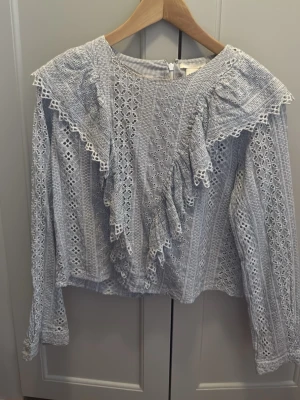 Blåvit blus med spets och volanger - Supersöt blåvit blus med tunna ränder, spetsdetaljer och volanger framtill och baktill. Blusen har långa ärmar och dragkedja i ryggen. Perfekt för dig som gillar romantisk och vintageinspirerad stil.