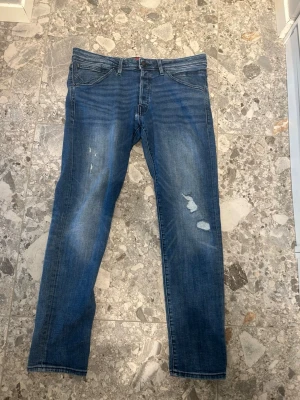 Blå slitna jeans med raka ben - Säljer ett par blå jeans med raka ben och slitningar på framsidan. Jeansen har klassiska fem fickor och en snygg tvättad look. Perfekta för dig som gillar en avslappnad och trendig stil.