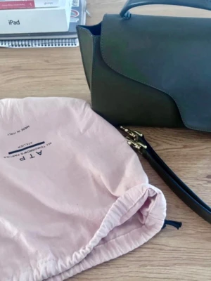 Grön handväska från ATP Atelier - Snygg grön handväska från ATP Atelier i slätt läder med unik asymmetrisk lockdesign. Väskan har ett topphandtag och avtagbar axelrem i svart läder med guldfärgade detaljer. Perfekt för dig som gillar stilrena och moderna accessoarer.