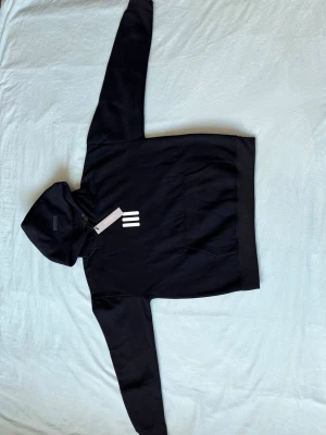 Essential svart hoodie med vit logga - Svart hoodie från Essential med vit minimalistisk logga på bröstet. Klassisk känguruficka framtill och huva med dragsko. Mjuk bomullskänsla och avslappnad passform, perfekt för en chill och stilren look.