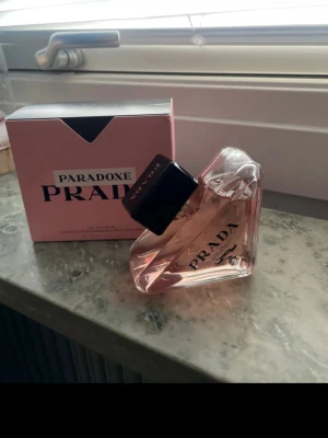 Prada Paradoxe Eau de Parfum - Prada Paradoxe Eau de Parfum med en unik, triangelformad flaska i rosa glas och svart kork. Doften kommer i en snygg rosa kartong med svart och vit detalj. Perfekt för dig som gillar exklusiva och moderna parfymer.