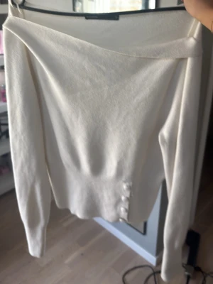 Off shoulder vit tröja med knappar - Trendig off shoulder tröja i vit färg med långa ärmar och dekorativa knappar vid sidan. Mjuk och stretchig kvalitet som sitter snyggt på kroppen. Perfekt för dig som vill ha en stilren men ändå unik look.