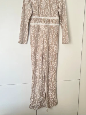 Lång beige spetsjumpsuit med blommönster - Elegant jumpsuit i beige med vit spets och blommigt mönster. Den har lång ärm, rund halsringning och fina spetsdetaljer i midjan. Perfekt för dig som vill ha en romantisk och stilren look.