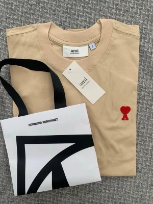 Beige t-shirt från Ami Paris - Snygg beige t-shirt från Ami Paris med klassisk passform och rund hals. T-shirten har Ami:s ikoniska röda hjärta broderat på bröstet. Tillverkad i mjuk bomull, perfekt för en stilren och avslappnad look.