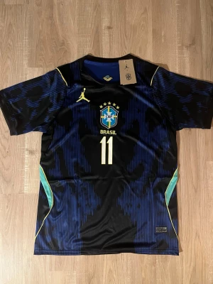 Brasilien Raphinha VM-2026 Fotbollströja  - Snygg svart och blå Brasilien fotbollströja med nummer 11 och Raphinha på ryggen. Tröjan har gula detaljer, CBF-emblem på bröstet och gröna inslag på sidorna. Perfekt för dig som älskar fotboll och vill sticka ut på planen. HELT NY