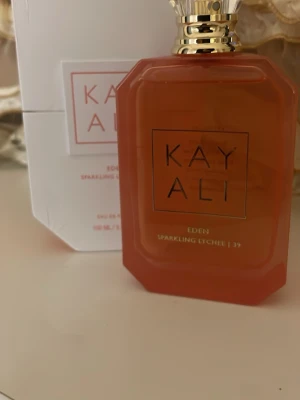 Kayali Eden Sparkling Lychee parfym - Kayali Eden Sparkling Lychee är en parfym i en snygg, rödorange, åttakantig glasflaska med guldfärgad kork. Doften har fruktiga och fräscha inslag av litchi. Flaskan har en modern design och kommer i en vit kartong med röd text.