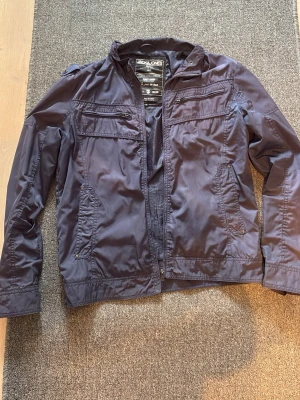 Mörkblå vindjacka från Jack & Jones - Snygg mörkblå vindjacka från Jack & Jones med flera dragkedjeförsedda fickor framtill och ståkrage. Jackan är tillverkad i ett lätt, glansigt material och har en modern, sportig look. Perfekt för dig som vill ha en stilren jacka med praktiska detaljer.