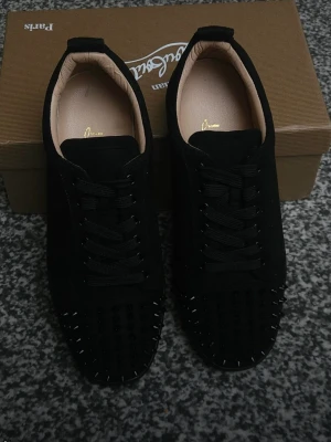 Svarta sneakers med nitar från Louboutin - Säljer ett par svarta sneakers från Christian Louboutin med coola svarta nitar på tån. Skorna har en klassisk låg siluett, svarta skosnören och ovandel i mocka. Insidan är ljusbeige och sulan är svart. Perfekta för dig som vill sticka ut med din stil.
