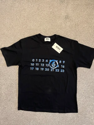Svart t-shirt från  Maison Margiela - Cool svart t-shirt från MM6 Maison Margiela med tryck av siffror i blått och vitt på bröstet där siffran 6 är markerad. Klassisk passform och rund halsringning. Perfekt för dig som gillar streetwear och vill sticka ut lite extra.