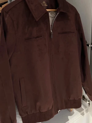 Brun mockajacka från Loro Piana - Snygg brun jacka i mocka från Loro Piana med dragkedja framtill, klassisk krage och ribbade muddar. Jackan har två bröstfickor med dragkedja och två sidofickor. Perfekt för dig som gillar stilrena och exklusiva plagg.