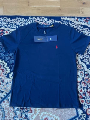 Mörkblå t-shirt från Polo Ralph Lauren - Klassisk mörkblå t-shirt från Polo Ralph Lauren med rund hals och ikonisk röd logga broderad på bröstet. Tillverkad i mjuk bomull och har en normal passform. Perfekt basplagg med stilren design.