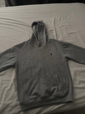 Grå hoodie från Polo Ralph Lauren - Snygg grå hoodie från Polo Ralph Lauren med klassisk broderad logga på bröstet. Hoodien har en stor känguruficka framtill och ribbade muddar vid ärmslut och nederkant. Perfekt för en chill och stilren look.