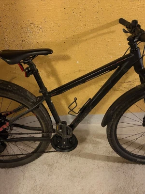 Svart mountainbike med skivbromsar - Säljer en svart mountainbike med grova däck, skivbromsar och stänkskärmar. Cykeln har flaskhållare och flera växlar, perfekt för både stad och terräng. Ramen är i metall och styret är rakt för bättre kontroll.