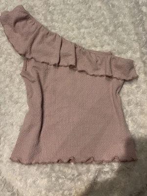 Ljusrosa oneshoulder topp med volang - Trendig ljusrosa topp i oneshoulder-modell med volang längs halsringningen. Toppens material har en lätt strukturerad känsla och den är perfekt för dig som vill sticka ut med en feminin och modern stil. Använd ca 2 gånger