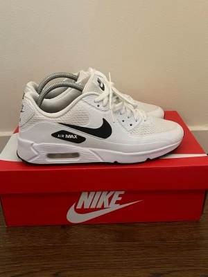 Nike Air Max vita sneakers - Nike Air Max sneakers i vitt med svart swoosh och Air Max-detalj på sidan. Ovandelen är i mesh och syntet för bra andningsförmåga och komfort. Klassisk låg modell med vit sula och svarta detaljer. Perfekta för dig som gillar stilrena och sportiga skor.