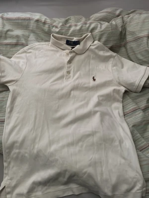 Vit pikétröja från Polo Ralph Lauren - Klassisk vit pikétröja från Polo Ralph Lauren med korta ärmar och krage. Tröjan har knappar framtill och den ikoniska broderade loggan på bröstet. Perfekt till jeans eller shorts för en clean och stilren look.