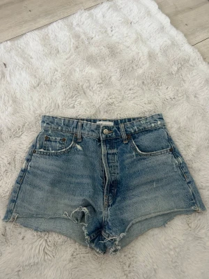 Blå denimshorts från Zara - Klassiska blå denimshorts från Zara med hög midja och råa kanter. Shortsen har fem fickor, knapp och dragkedja framtill samt en avslappnad passform. Perfekta för varma dagar och enkel att matcha med olika toppar.