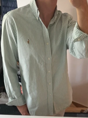 Grön- och vitrandig skjorta Polo Ralph Lauren - Klassisk grön- och vitrandig skjorta från Polo Ralph Lauren med button-down krage och broderad logga på bröstet. Skjortan har långa ärmar och knäppning framtill. Perfekt för en stilren och avslappnad look.