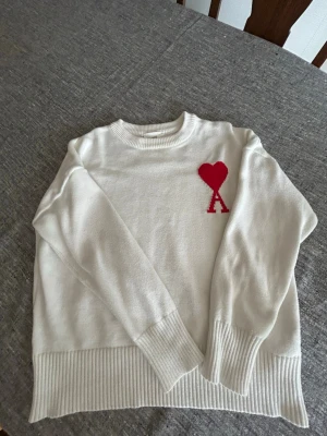 Vit stickad tröja från Ami - Vit stickad tröja från Ami med ribbade muddar och nederkant. På bröstet finns ett rött hjärta och bokstaven A broderat. Tröjan har rund halsringning och är gjord i mjukt material, perfekt för en stilren och trendig look.