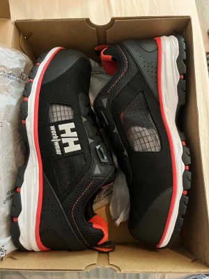 Chelsea Evo 2 Sandal BOA S1P - Säljer ett par helt oanvända svarta Helly Hansen Chelsea Evo 2 Sandal BOA S1P med röd och vit sula. Skorna har meshpaneler, BOA-snörning och komposittåhätta. Perfekta för dig som vill ha slitstarka och ventilerande arbetsskor med sportig look. 