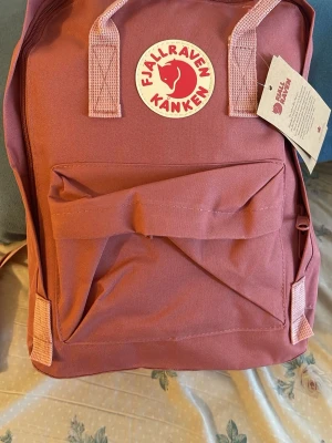 Rosa Kånken ryggsäck från Fjällräven - Säljer en klassisk rosa Fjällräven Kånken ryggsäck med det ikoniska runda logotypmärket framtill. Ryggsäcken har ett stort huvudfack, en framficka med dragkedja och sidofickor. Tillverkad i slitstarkt syntetmaterial med matchande handtag och axelband.