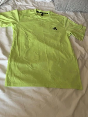 Grön Adidas t-shirt i funktionsmaterial - Fräsch gul t-shirt från Adidas med klassisk logga på bröstet. Tillverkad i ett lätt och ventilerande funktionsmaterial, perfekt för träning. T-shirten har rund halsringning och korta ärmar för en sportig look.