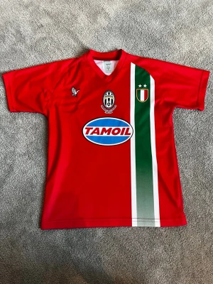 Juventus 2004/05 away shirt Zlatan Ibrahimovic - Röd Juventus matchtröja med grönt och vitt band på framsidan, Juventus märke, Tamoil-logga och nummer 9 med Ibrahimovic på ryggen. Korta ärmar och perfekt för fotbollsfans som vill sticka ut på planen eller läktaren med en klassisk retro tröja.