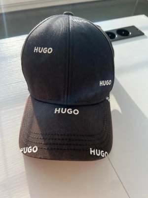 Svart keps från HUGO med logga - Svart keps från HUGO med vit logotyp tryckt flera gånger över hela kepsen. Justerbar rem baktill med röd detalj och HUGO-tryck. Klassisk böjd skärm och sex paneler. Tillverkad i bomullstyg.