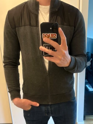 Mörkgrå jacka/cardigan från COLMAR - Snygg mörkgrå jacka från Colmar med svarta detaljer över axlarna. Jackan har ribbade muddar, hög krage och dragkedja framtill. Två fickor med dragkedja och ett Colmar-märke på ärmen. Perfekt för en stilren och avslappnad look. 