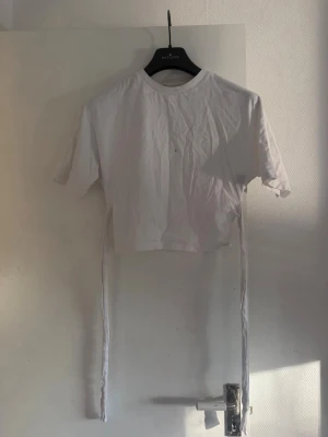 Vit croppad topp med knyt från Nelly - Vit croppad topp från Nelly med korta ärmar och rund halsringning. Toppen har långa knytband i sidorna som ger en trendig look. Perfekt att styla med jeans eller kjol för en cool vibe. Har sminkfläck 