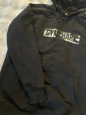 Svart Gymshark hoodie med camologga - Svart hoodie från Gymshark med en fet camoinspirerad logga på bröstet. Klassisk känguruficka framtill, ribbade muddar och justerbar huva med snören. Perfekt för chill eller gymmet. Mjuk bomullsblandning som känns skön mot huden.