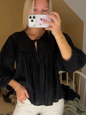 Svart blus med knytband - Svart blus med knappar framtill och knytband vid halsen. Blusen har en lös passform och trekvartsärmar med lätt puff. Tunt, flowigt tyg som ger en avslappnad och trendig look.