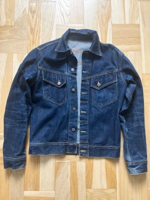 Mörkblå jeansjacka från Nudie Jeans - Snygg mörkblå jeansjacka från Nudie Jeans med klassisk passform, bröstfickor med lock och knappar framtill. Jackan har kontrastsömmar och slitningar för en cool look. Perfekt att slänga över en hoodie eller t-shirt.