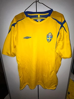 Svensk landslagströja Umbro gul/blå - Gul fotbollströja från Umbro med blå detaljer och Sveriges landslagsemblem på bröstet. Från 2006 i storlek L. Klassisk rund halsringning och korta ärmar. Perfekt för dig som vill visa ditt stöd för Sverige nu i VM.