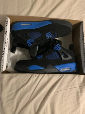 Air Jordan 4 Retro Black/Blue - Snygga Air Jordan 4 Retro sneakers i svart och blått med klassisk siluett. Ovandel i mocka och mesh, detaljer i blått och svart plast samt synlig Air-enhet i sulan. Perfekta för dig som vill sticka ut med en ikonisk och sportig look.