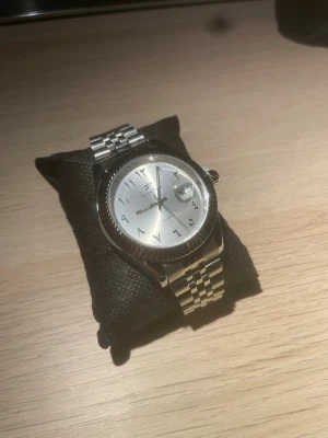 Silverfärgad klocka med arabiska siffror - Stilren klocka från Ricecgo med silverfärgad urtavla och arabiska siffror. Klockan har en klassisk länk i metall och datumvisning vid klockan tre. Perfekt accessoar för dig som gillar en clean och tidlös look.