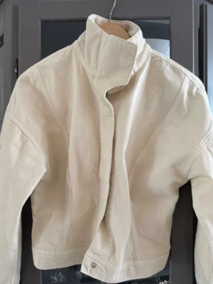 Beige croppad jacka med hög krage - Trendig beige jacka med croppad passform och hög krage. Jackan har långa ärmar och stilren design utan synliga detaljer. Perfekt för dig som gillar minimalistisk stil och vill ha något som sticker ut.