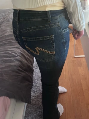 Mörkblå bootcut jeans - Säljer ett par mörkblå lågmidjade jeans med detaljer på fickorna i beige. Jeansen har klassisk femficksdesign. Perfekta för dig som gillar en lite mer unik detalj på dina jeans. Jeansen har tagits bort från hemsidan. Nypris 700kr