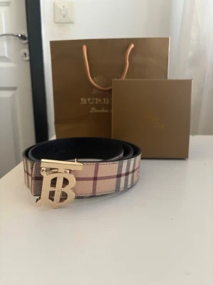 Burberry rutigt bälte med gulddetalj - Snyggt bälte från Burberry med klassiskt rutigt mönster i beige, rött och svart. Bältet har ett stilrent guldfärgat spänne med Burberrys ikoniska logga. Perfekt accessoar för att lyfta din outfit.