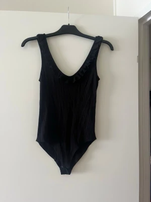 Svart ribbad linnetopp med volang - Stilren svart body med ribbad struktur och djup urringning. Volangdetalj längs halsringningen ger en extra touch. Ärmlös modell i stretchigt material som sitter snyggt på kroppen.