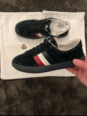 Moncler svarta sneakers med röd/vit detalj - Snygga svarta sneakers från Moncler i mocka med klassiska röd och vita ränder på sidan och Moncler-logga. Skorna har svarta snören, svart sula och vit insida. Perfekta för dig som gillar stilrena och exklusiva sneakers med en sportig touch. sulan är ganska sliten dustbag kommer ej